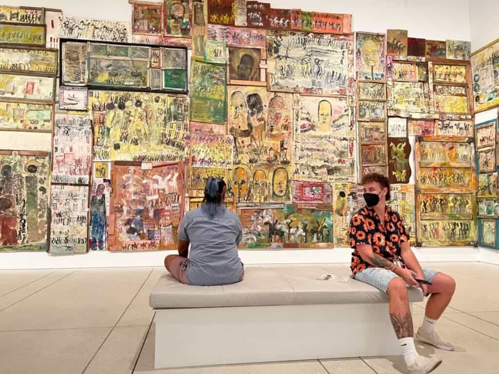 Two transmasculine people sit on a white bench. One faces away from the camera and toward a wall completely covered in small