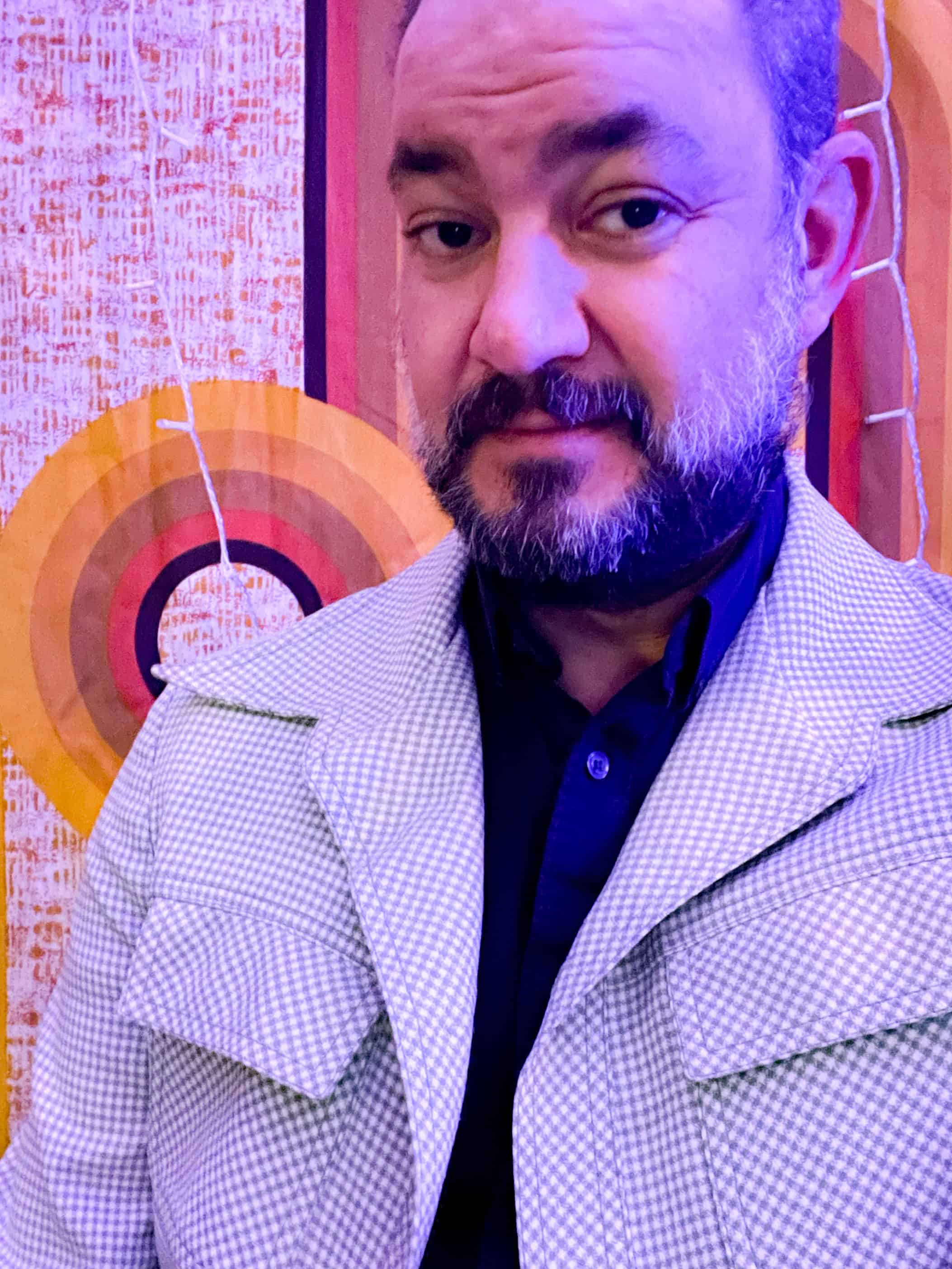 Aaron has dark facial hair and fair skin. He wears a blue button down, a tan jacket, and stands in front of a multicolored background.