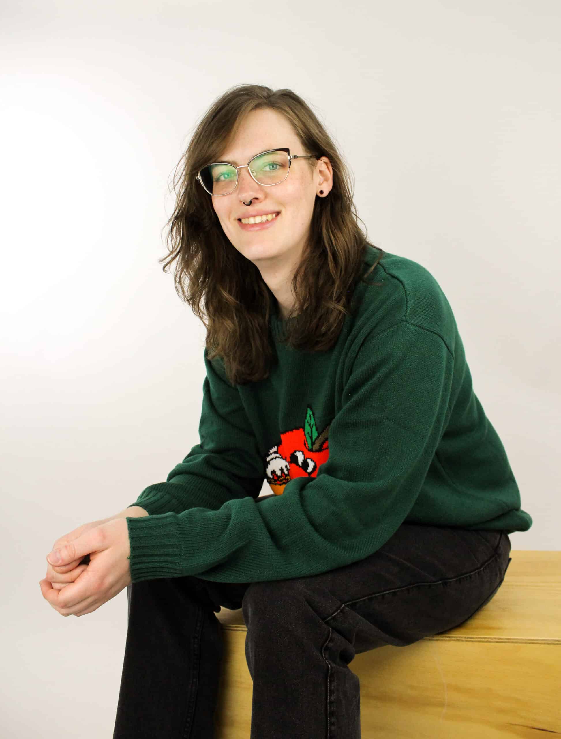Sagen wears black jeans and a dark green sweater with red embroidery. She smiles at the camera. She has wavy brown shoulder length hair, glasses, and a black septum ring. 