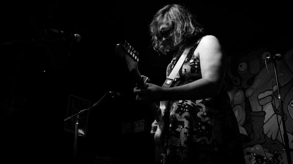 Maggie plays guitar on stage while wearing a floral dress. The photo is in Black and white.|||Aaron has dark facial hair and fair skin. He wears a blue button down