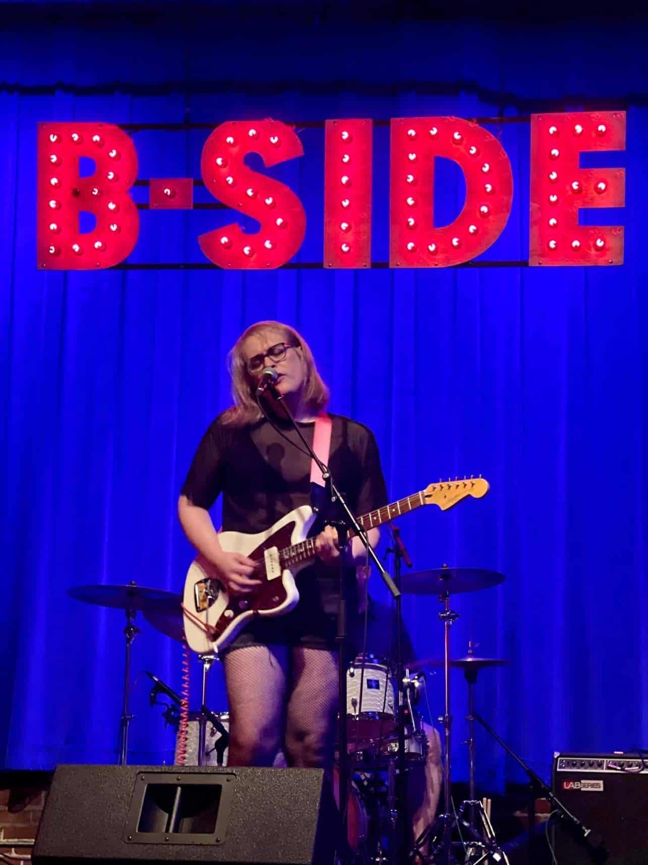 Maggie, blonde with black frame glasses, sings into the mic with her eyes closed. She plays a white electric guitar and wears a black dress. She is standing on stage in front of a blue curtain with the Word "B-Side" hanging in red at the top. 