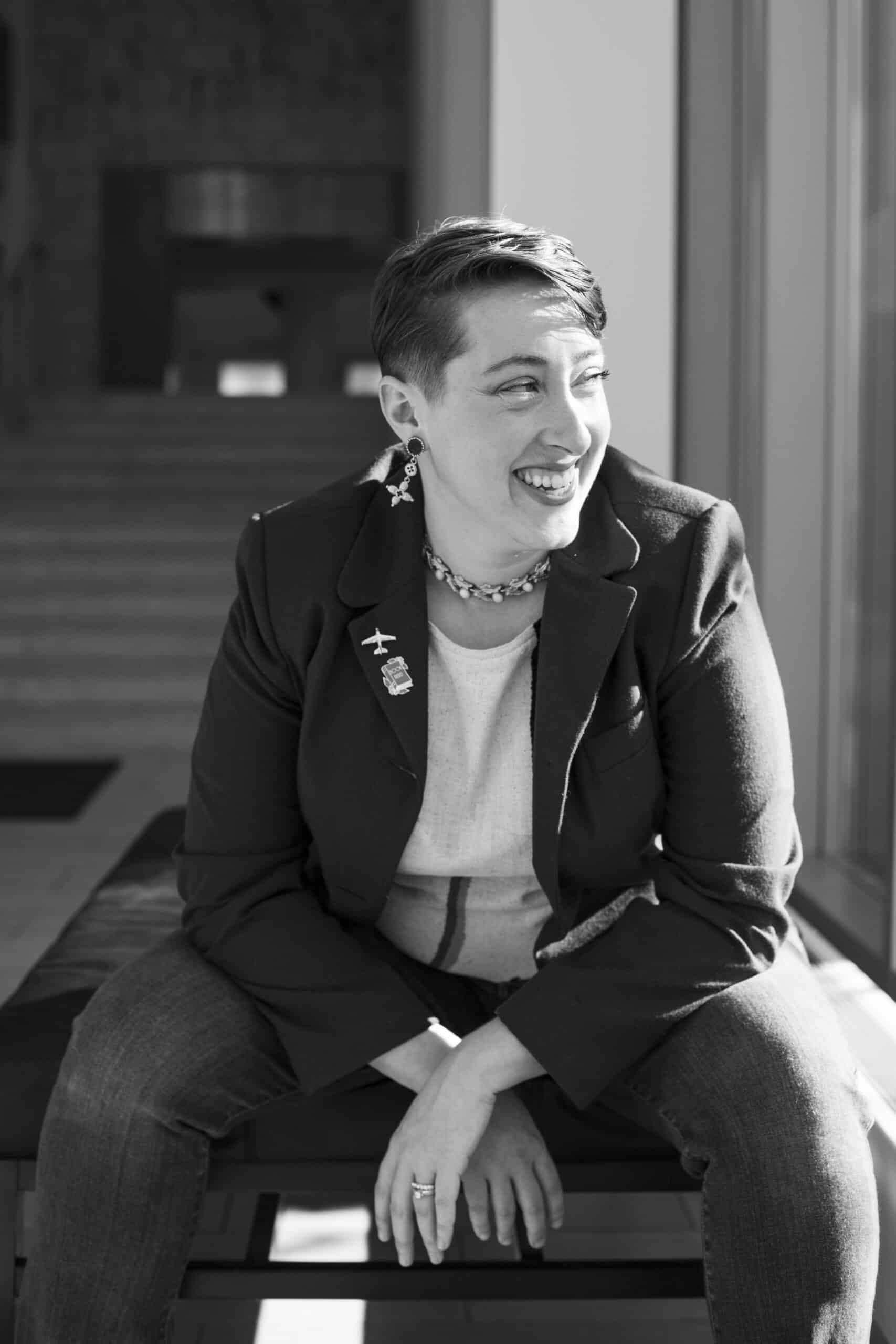Mandy has short hair, dangling earrings and fair skin. They wear a blazer and look away from the camera while laughing. The photo is in black and white. 