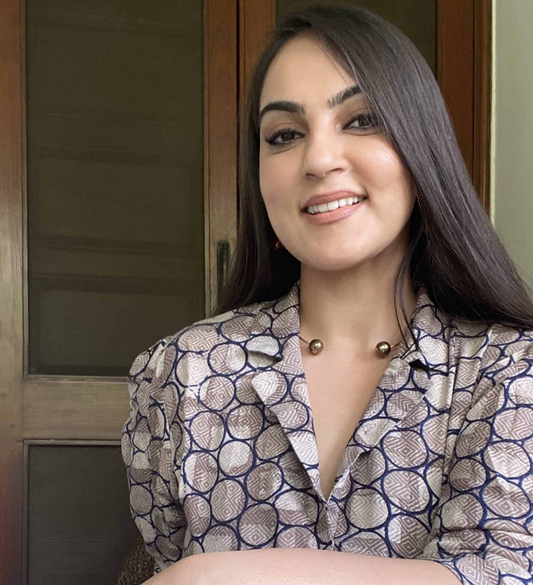 Noor has long dark brown hair and fair skin. She smiles at the camera in front of wooden doors and wears a neutral printed shirt. 