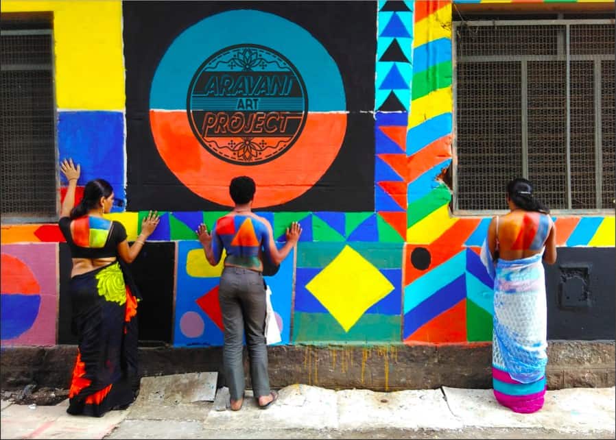Women stand in front of a colorful mural with their backs facing the camera. Part of their backs are painted to blend in with the mural. Above their head there is a painted logo for the Aravani Art Project. 