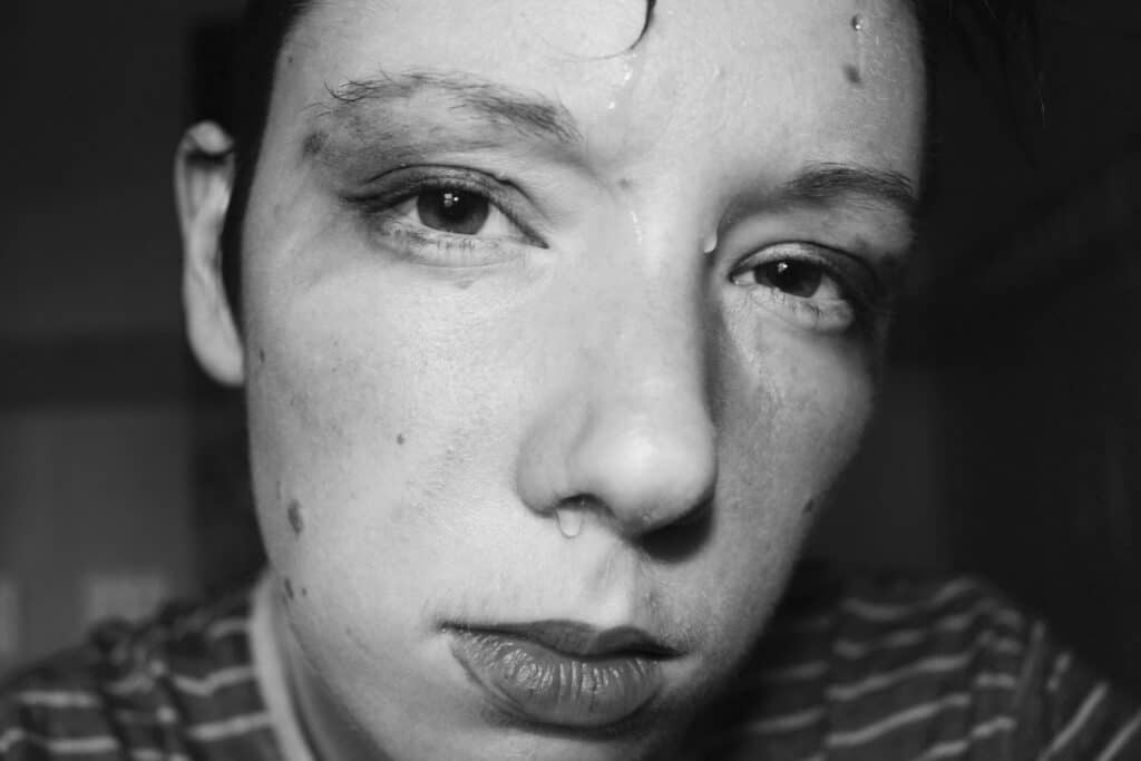 Lexie Bean stares directly into the lens and places their face close to the camera. The photo is black and white. They have sweat dripping down their face and some darker substance smeared above their eyes.|Lexie Bean is fair skinned and wears a burgundy pattered button down shirt. He has short