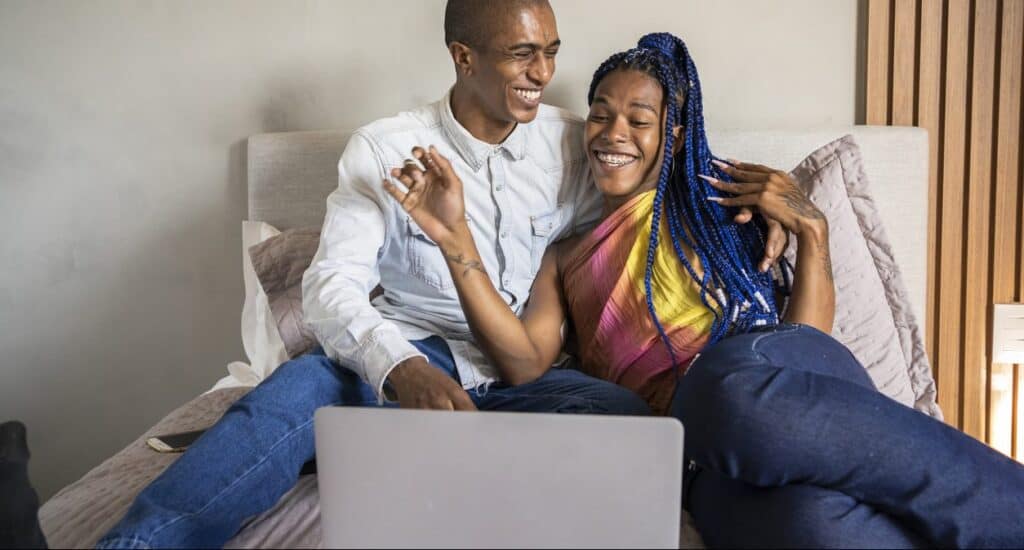 A Black LGBT couple watches a movie on a laptop in bed while laughing. One person wears a colorful top with blue box braids. The other wears a light blue button up and has a shaved head.|