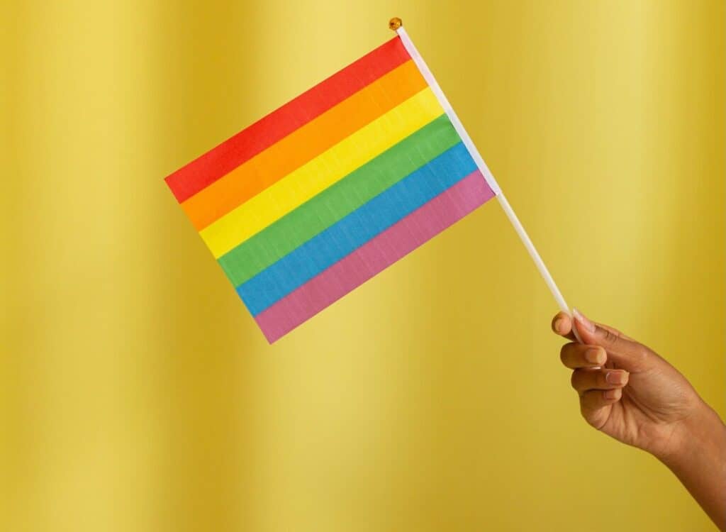 A brown hand holds a pride flag against a yellow background.||