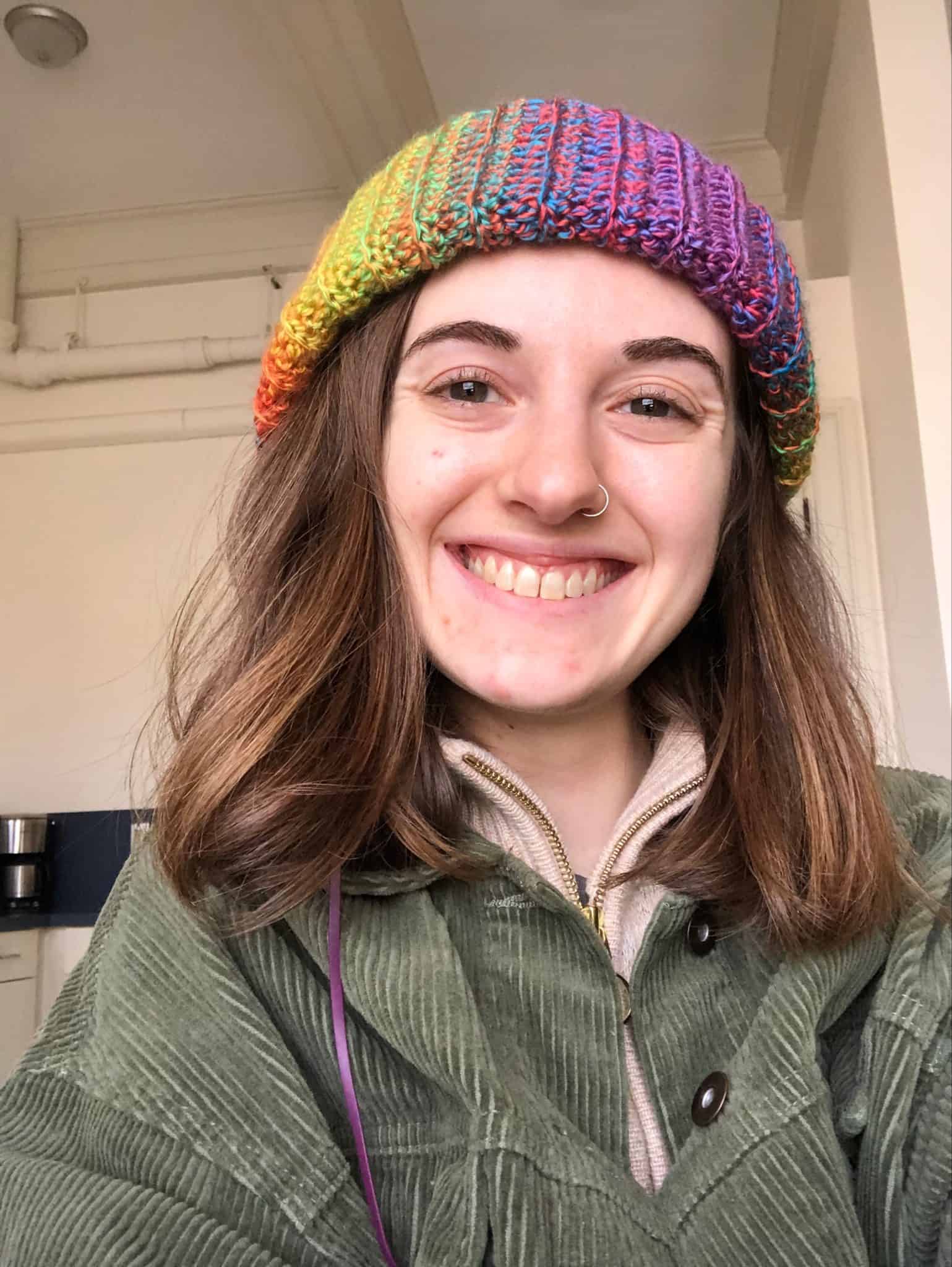 Maya smiles at the camera. She has straight brown shoulder-length hair. She wear a rainbow knitted beanie and a forest green jacket. 