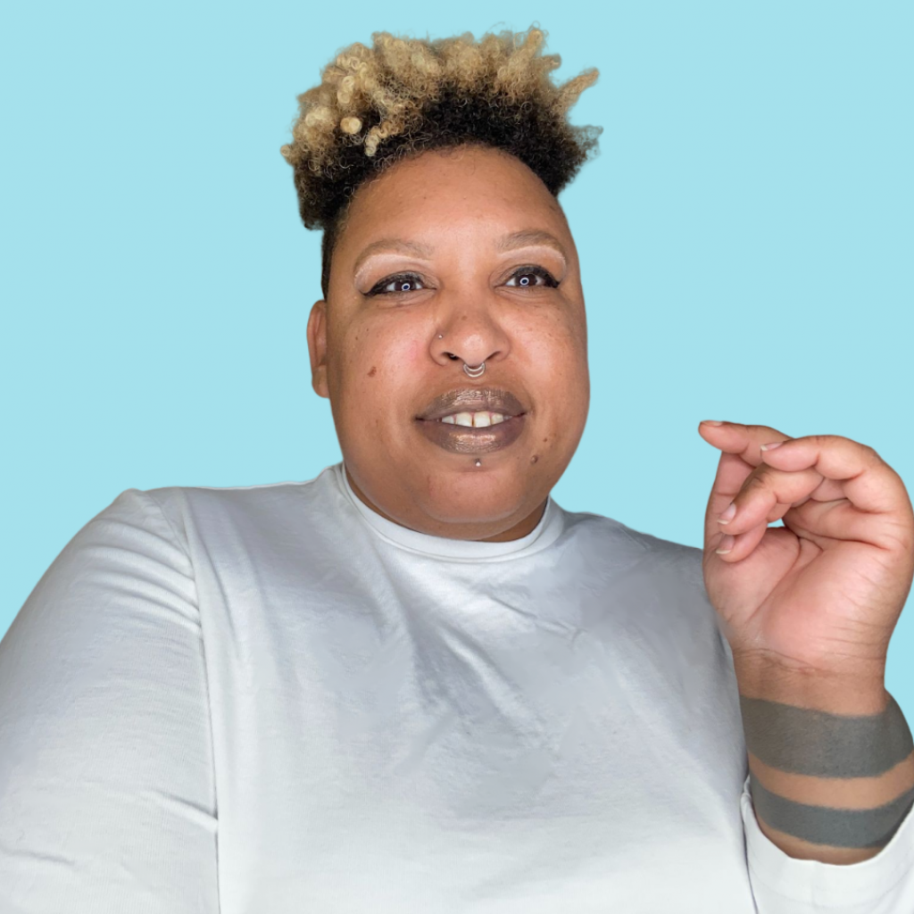 Image description: Head and shoulders shot of Dominic against a light blue background. Dominic is a Black non-binary person wearing a white crop top. Dominic sports short curly hair with blonde tips and light makeup. Dominic is smiling into the camera with one hand in a casual pose.