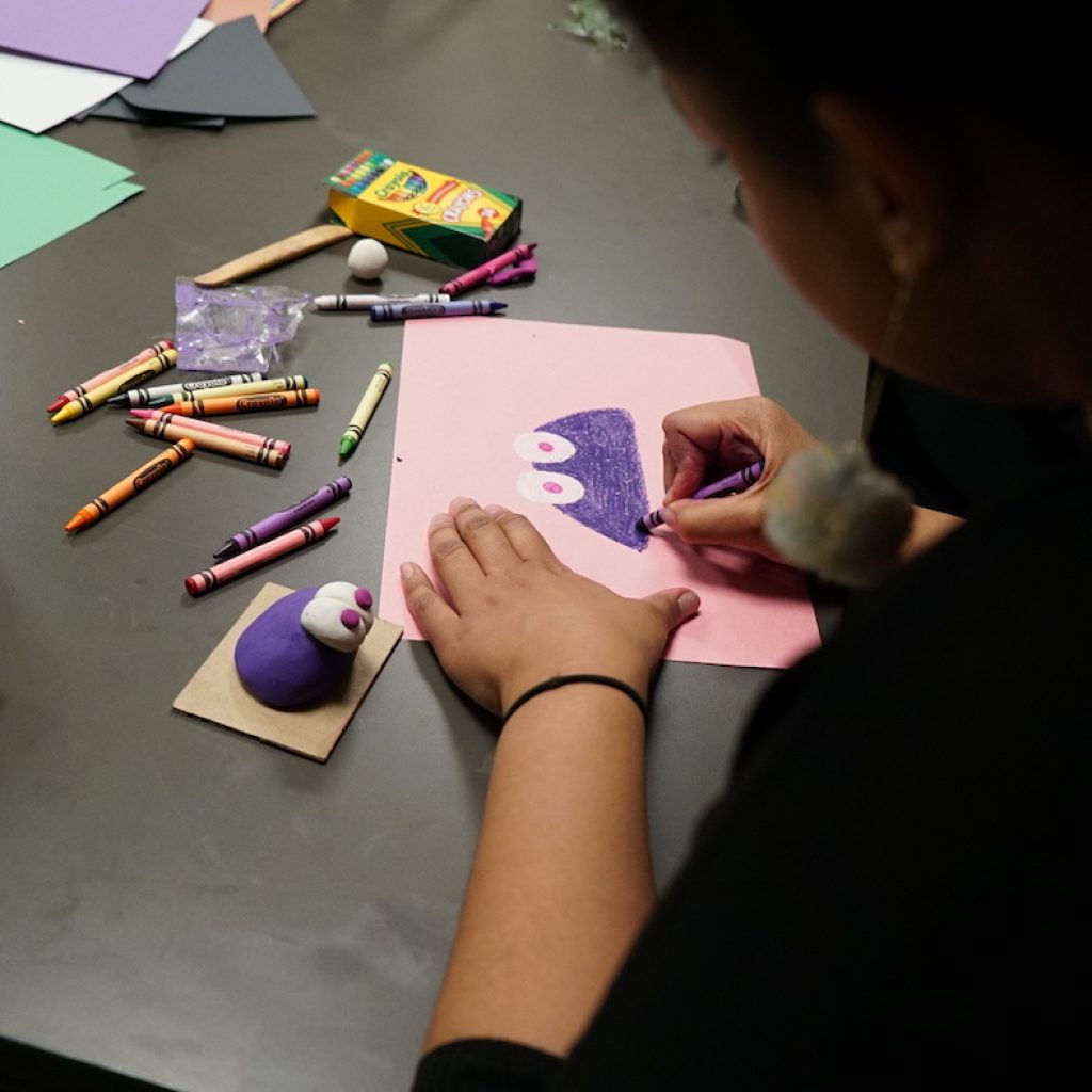 A fair skinned brown person leans over a drawing. On a pink piece of paper their are drawing a purple figure in the shape of a half-circle with big eyes and red pupils. Crayons are splayed in front of them on the table. To their left, the purple figure in clay form sits on top of a small piece of cardboard. 