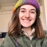 Maya smiles at the camera. She has straight brown shoulder-length hair. She wear a rainbow knitted beanie and a forest green jacket. 