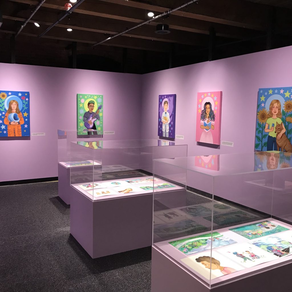 Noah's portraits hang on purple walls. Glass covered podiums containing smaller drawings are in the middle of the room. 