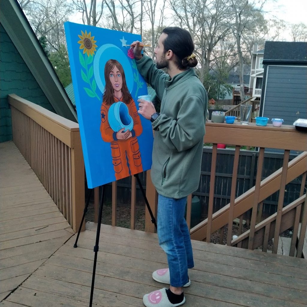 Noah Grigini paints a bright blue portrait of Olly as an astronaut while standing outside on a wooden deck. Grigini wears a green jacket, jeans, and slippers with their hair pulled back in a ponytail.