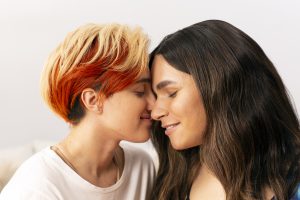Two trans people lovingly resting their faces against each others. The fair skinned person on the left wears a white t-shirt and has short ombre blonde and red hair. The fair-skiiner person on the right has long brown hair and wears a black t-shirt.