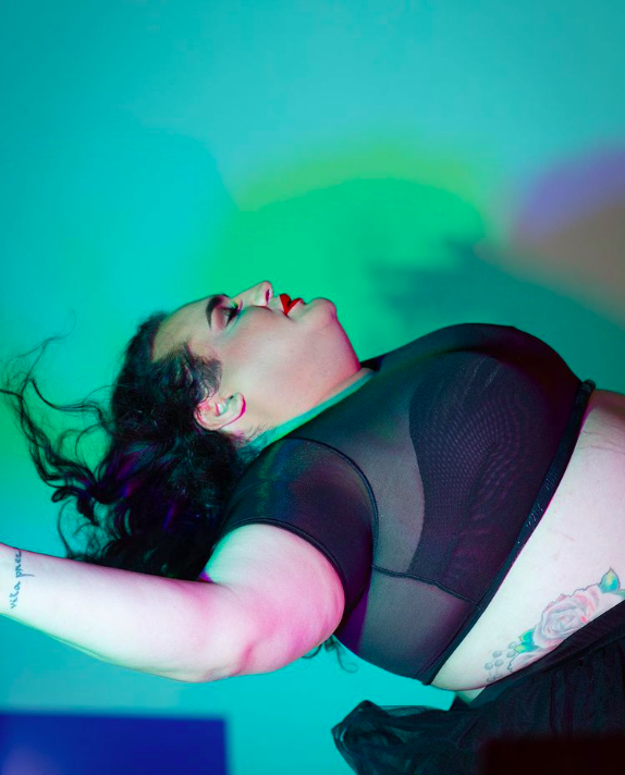 Jayla bends backwards in a sheer black crop top against a green and blue background. She wears red lipstick and her black hair trails behind her.