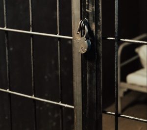 The corner of a jail cell with the corner of a bed with white sheets, metal bars, and an open padlock.