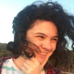 Alex has long curly black hair that is flowing in the wind. They are fair skinned, smiling, and looking off to the side while holding their chin.