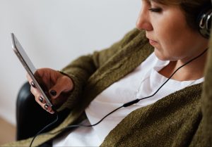 A person with black painted fingernails, short brunette hair, and a green sweater looks at their iphone while wearing headphones.