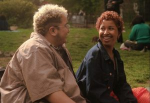 Two trans people with short curly hair smiling and looking at each other while sitting in the park.