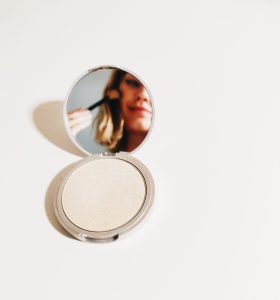 a person looks into a small ahnd mirror while brushing blush across their cheek. the background is white.