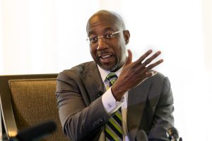 Senator Raphael Warnock is wearing a brown suit, green tie, silver rimm glasses, and sitting in a brown chair. He is waving a hand while he speaks. He has brown skin and a goatee.