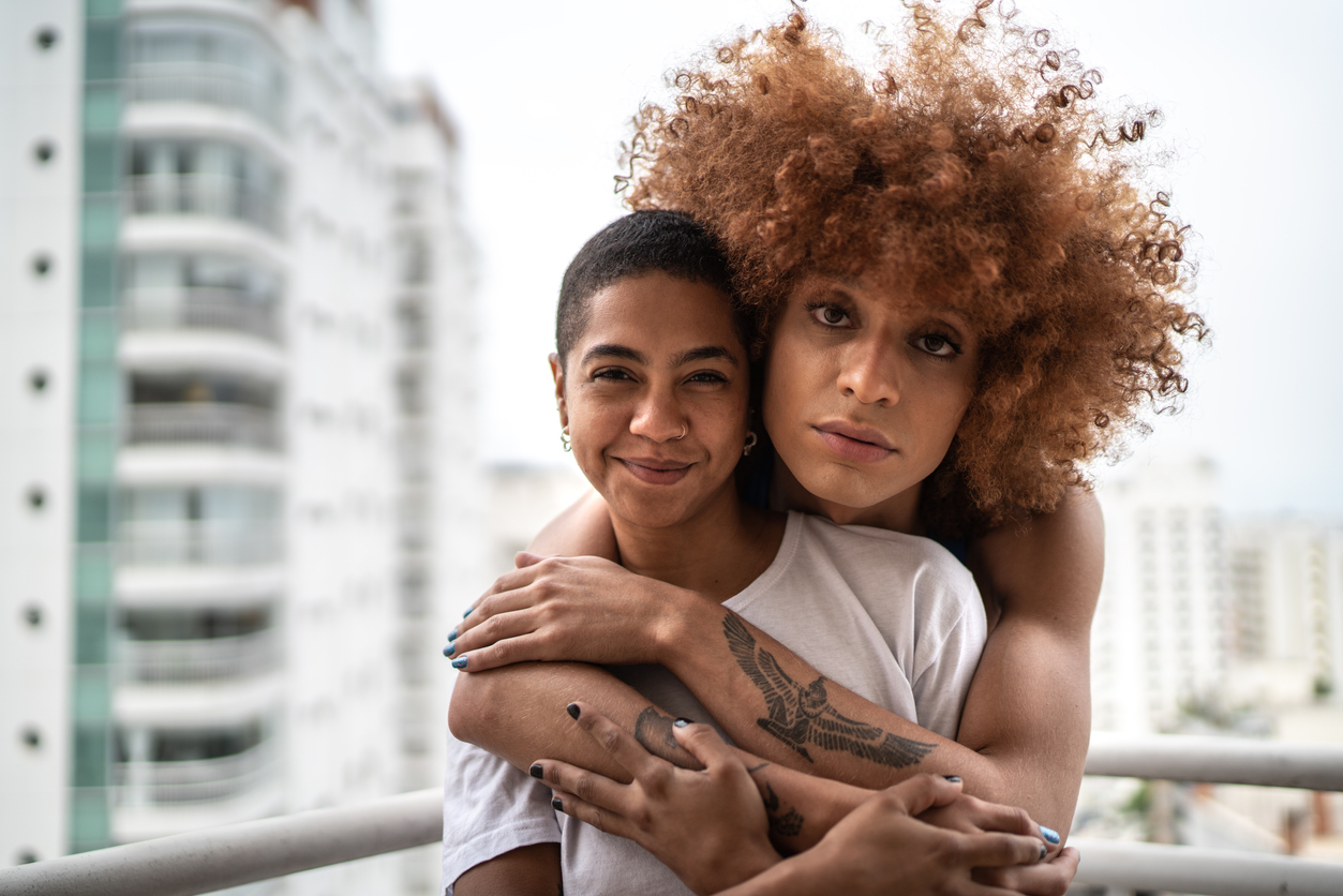 Two brownskinned people face the camera. The one in the back has a big copper colored afro and wraps their arms around the person in the front. The person in the front smiles, has short black hair, and wears a white tshirt.