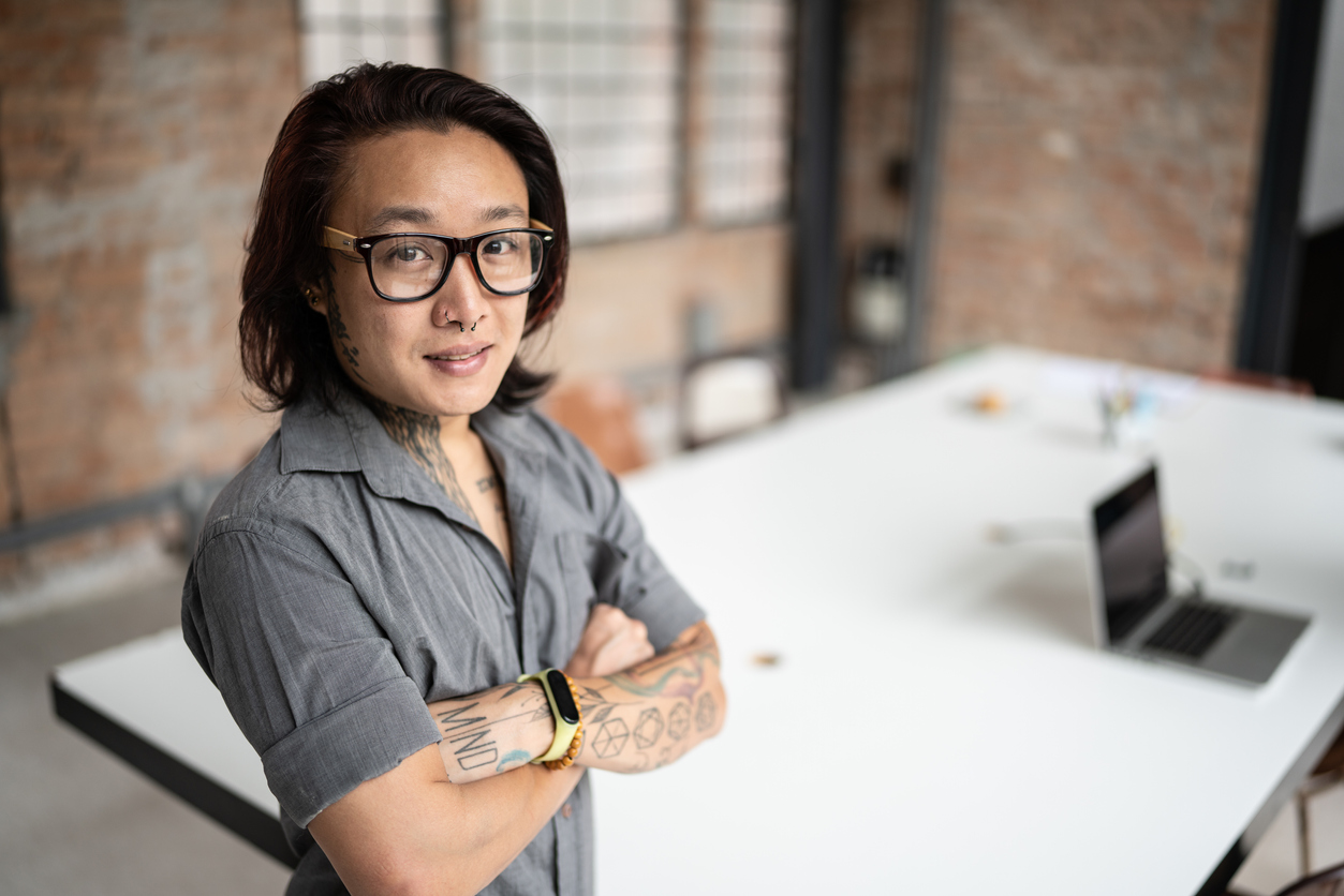 An asian transgender man stands confidently with his arms crossed in the left corner of the screen. He wears a grey short sleeve button down. In the background there is an office table in a brick room.