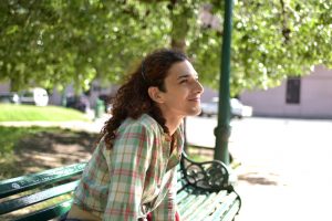 A trans woman sits on a green bench in a park. She is facing away from the camera so we see her profile. She has brown curly hair, wears a green flannel, and smiles while looking up.