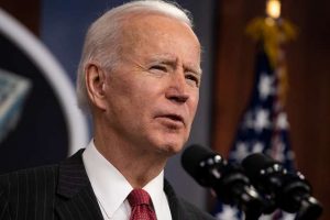 President Joe Biden speaking in front of a mic. He wears a black suit, white shirt, and red tie. A piece of the American flag can be seen in the background.