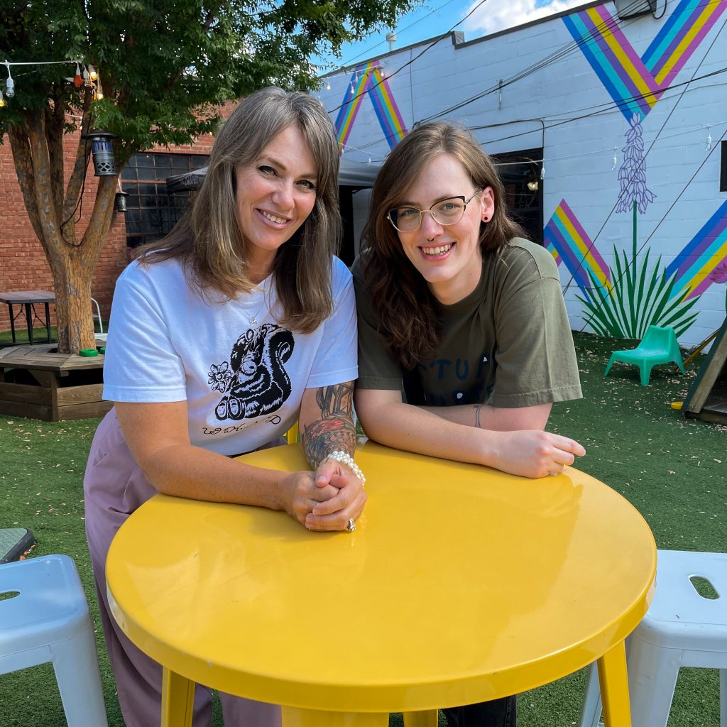 Victoria Cocklin (Left) leans on a yellow table and smiles at the camera. She has shoulder length hair, a white tshirt, and pink pants. Sagen Cocklin (Right) leans on the yellow table. They wear a dark green shirt and smile directly at the camera. Both Victoria and Sagen are in an outdoor lounge area with colorful triangles painted on a wall nearby. 
