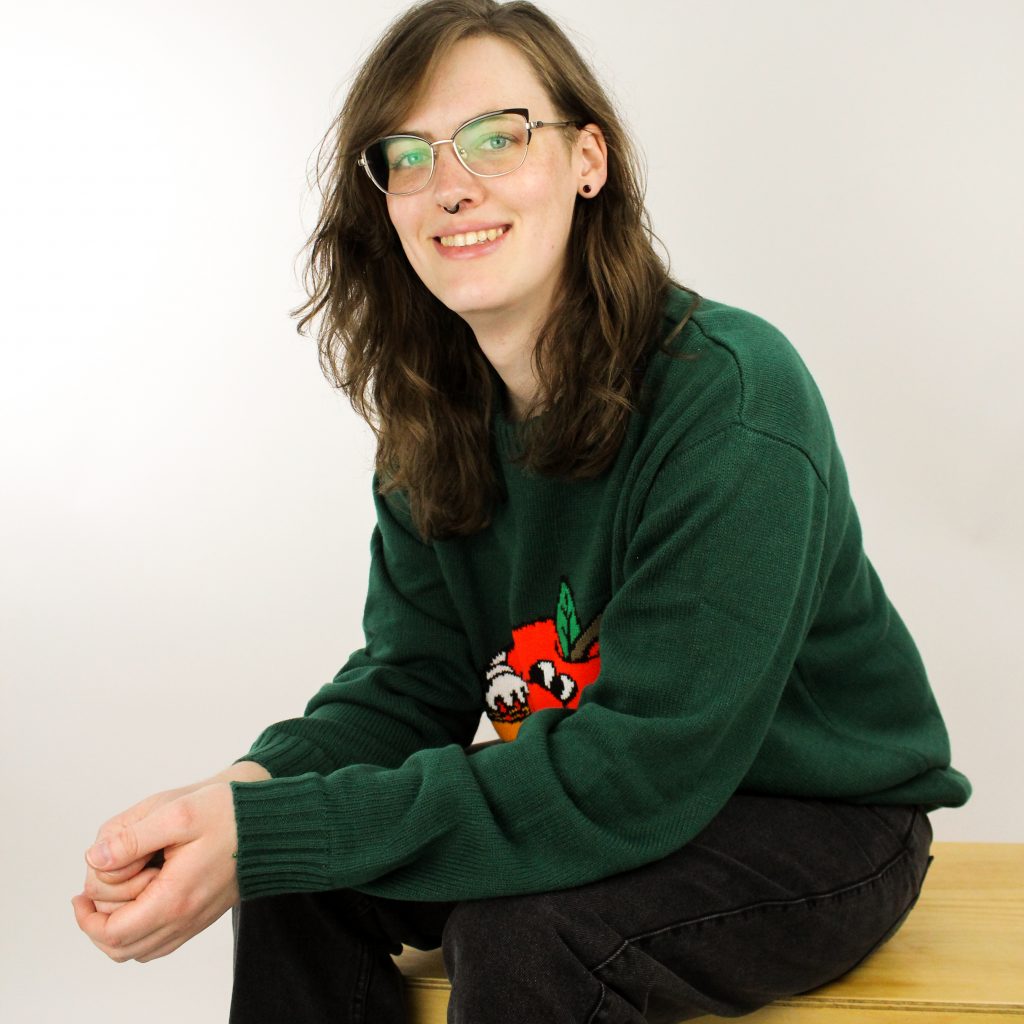 Sagen wears black jeans and a dark green sweater with red embroidery. She smiles at the camera. She has wavy brown shoulder length hair, glasses, and a black septum ring. 