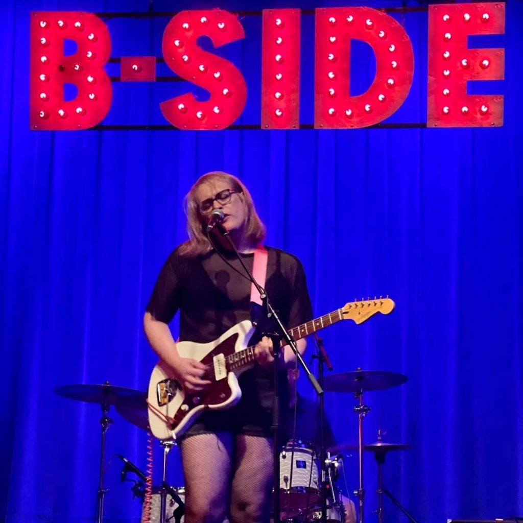 Maggie, blonde with black frame glasses, sings into the mic with her eyes closed. She plays a white electric guitar and wears a black dress. She is standing on stage in front of a blue curtain with the Word "B-Side" hanging in red at the top. 