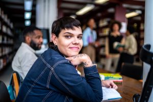 Portrait of a young transgender woman in a library with other people. They are wearing a blue jean jacket and looking over their shoulder at the camera. They have fair skin, black hair with fringe, and tattoos.
