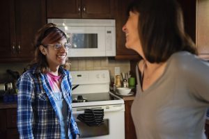 A trans teen with glasses, a blue flannel and brown skin smiles at their guardian while standing in the kitchen.