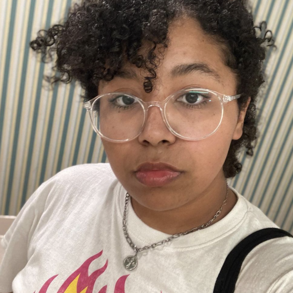 BMUD ANGEL in a selfie, they are wearing glasses and have their curly hair parted in a bang to the side with a stern look on their face