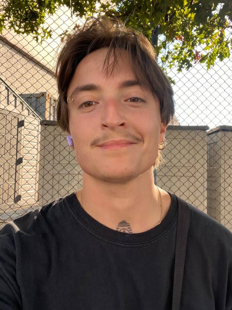 A young, white transmasculine person stands in front of a chain link fence. They have short brown hair, a