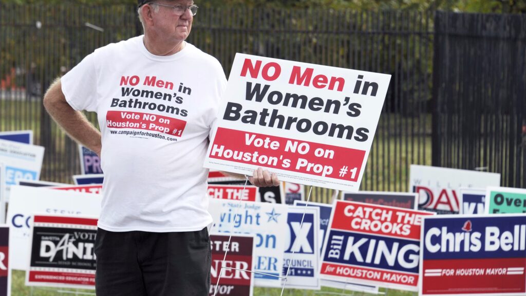 An older white man with gray hair and glasses stands in front of over 20 political lawn signs advertising candidates for Houston mayor. He is holding a sign that reads "NO Men in Women's Bathrooms. Vote NO on Houston's Prop #1" in red, black, and white print. His white T-shirt displays the same text as the sign he is holding.