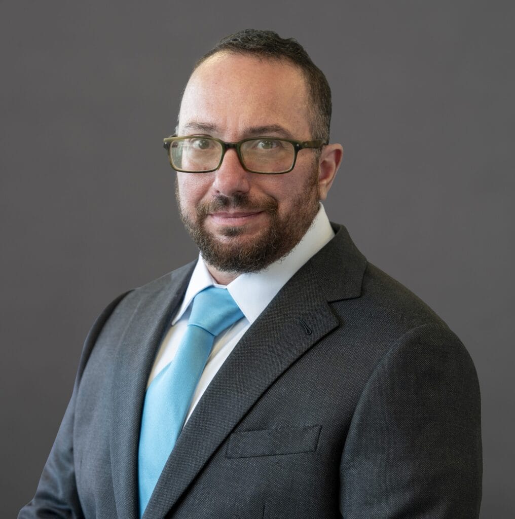 A headshot of Evan against a gray background. He wears rectangular glasses, a short beard, and a business suit with a turquoise tie.