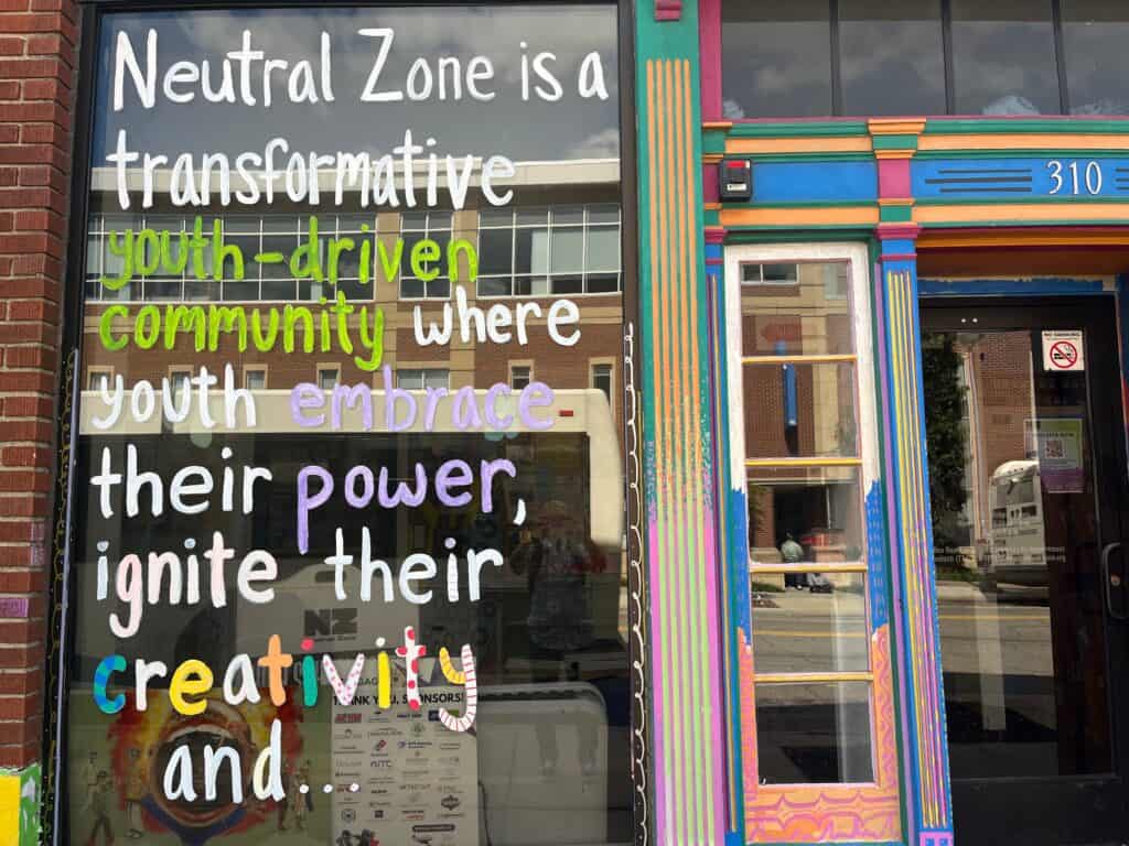 The colorful exterior doorway and front window of Neutral Zone. The window is painted with a quote: "Neutral Zone is a transformative youth-driven community where youth embrace their power, ignite their creativity and..."