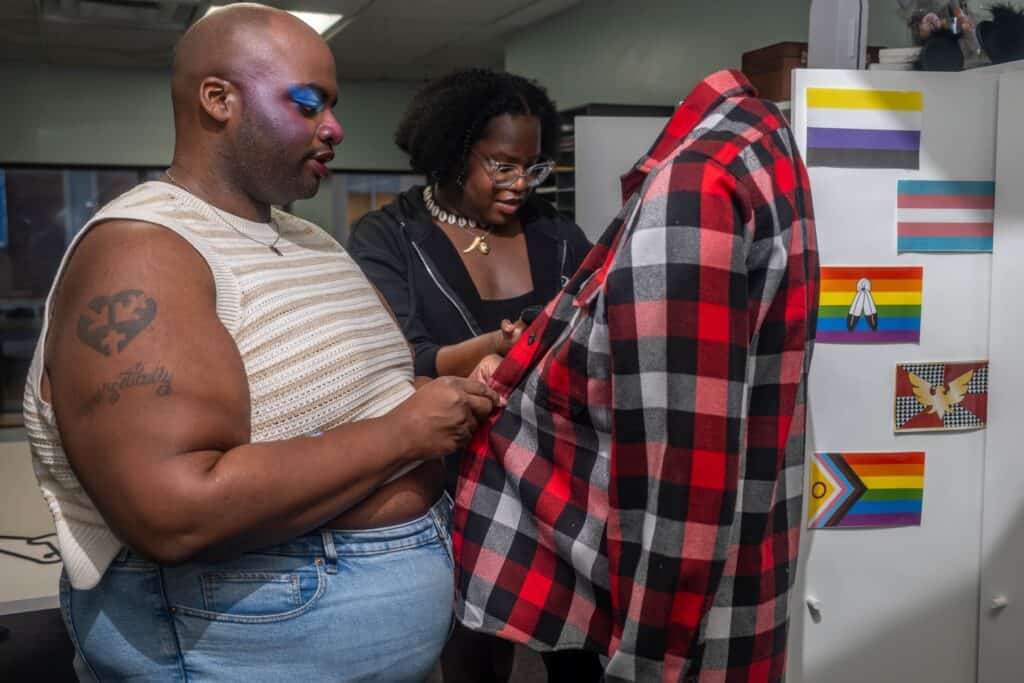 Two Black young adults stand in front of an open white closet with multiple LGBTQ+ flags pasted onto it. They are buttoning a red-and-black flannel shirt on a mannequin.