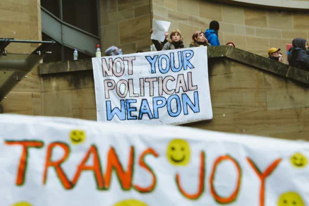 On a staircase, people hold up a sign that reads, "NOT YOUR POLITICAL WEAPON" in pastel pink and blue letters. In the foreground, a banner with smiley faces on it reads, "TRANS JOY" in orange ink.