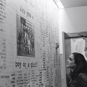Arielle Rebekah looks up at a wall covered in words and art. One part of the wawll reads "Are you a boy or a girl?" They have dark hair past their shoulders and are wearing a button down. The photo is in black and white.