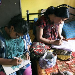 Two women sit next to each other while drawing. THey are wearing colorful garments and sitting in a colorful room