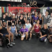 A group of queer and trans folks of varying sizes, shapes, racial identities, and abilities huddle together and smile at the camera.