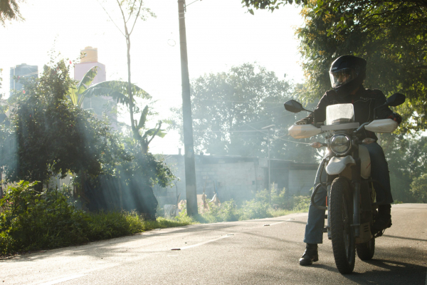 Yams, dressed in all black motorcycle gear, gets ready to ride. Yams is on the road with a luscious green background.