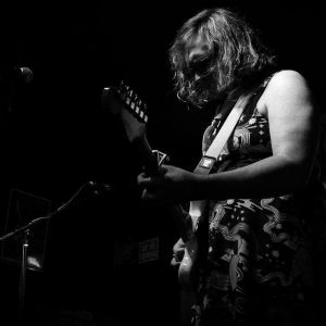 Maggie plays guitar on stage while wearing a floral dress. The photo is in Black and white.