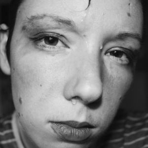 Lexie Bean stares directly into the lens and places their face close to the camera. The photo is black and white. They have sweat dripping down their face and some darker substance smeared above their eyes.