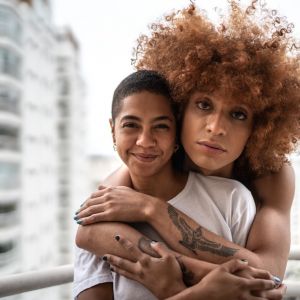 Two brownskinned people face the camera. The one in the back has a big copper colored afro and wraps their arms around the person in the front. The person in the front smiles, has short black hair, and wears a white tshirt.