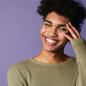 a genderqueer person with tan skins and curly black hair smiles in front of a purple background. They have glittery eye makeup.