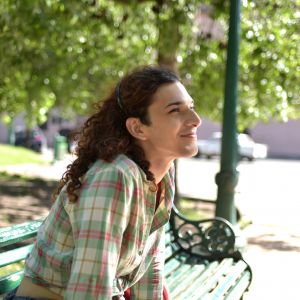 A trans woman sits on a green bench in a park. She is facing away from the camera so we see her profile. She has brown curly hair, wears a green flannel, and smiles while looking up.